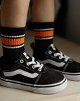 Child's legs wearing black sneakers with white laces, black and orange socks, and checkered shorts.