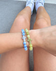 Restoration Cross yellow beaded bracelet styled with a blue bracelet on a wrist above crossed legs