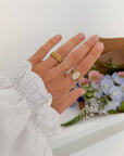 Hand wearing the Bloom Again Enamel Ring with other gold-tone rings beside pastel flowers