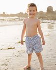 Young boy standing on a beach wearing patterned shorts.