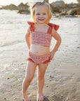Young girl in a red checkered swimsuit standing on a sandy beach with rocks and water in the background.