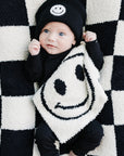 Baby wearing a black outfit with smiley face design, lying on a black and white checkered blanket.