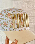 Floral-patterned cap with gold letters held by a hand against a tiled wall.