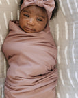 Newborn baby wrapped in a pink blanket with a matching headband on a beige and white patterned background