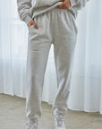 Person wearing a gray sweatshirt and pants with white sneakers in front of a white curtain.