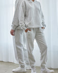 Two people wearing matching gray tracksuits standing in a bright room with white curtains.