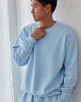 Person wearing a light blue sweatshirt and pants set against a white curtain.