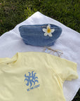 Morning Sun Be the Light tee laid flat with a small blue chest graphic beside a denim cap