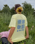 Model seated among wildflowers wearing the Morning Sun Be the Light tee with its back graphic visible