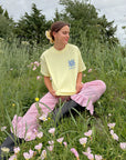 Model seated in a grassy field wearing the Morning Sun Be the Light tee with pink gingham pants