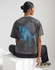 Model seated wearing the Lamp Black Believe in Miracles Heavyweight Tee with a large blue water-inspired back graphic