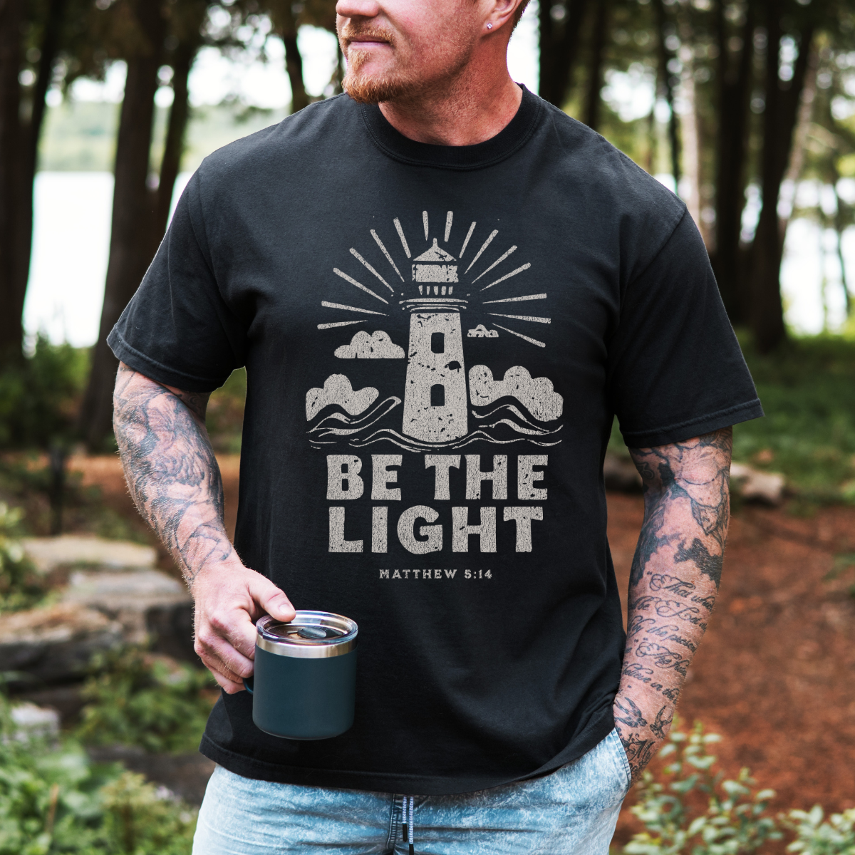 Man wearing a black t-shirt with a lighthouse design and 'Be the Light' text, holding a mug outdoors.