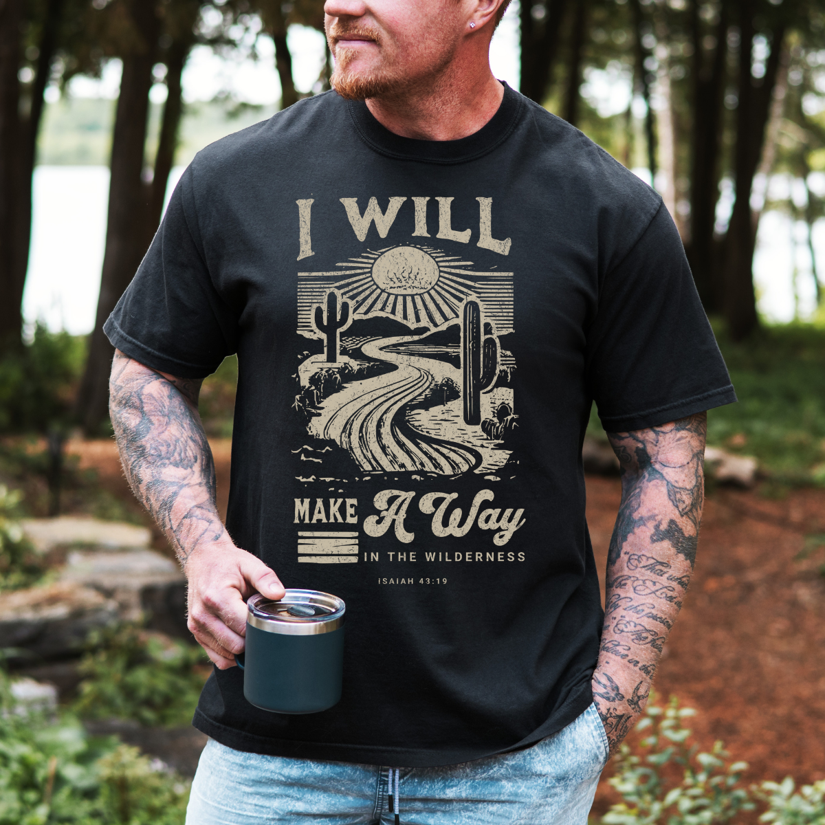 Man wearing a black t-shirt with a desert graphic and text, holding a mug outdoors.