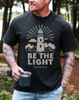 Man wearing a black t-shirt with a lighthouse design and 'Be the Light' text, holding a mug outdoors.