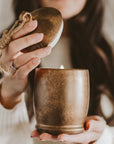 Holiday soy candle in an antique gold ceramic bell vessel, held with its matching rope-accented lid lifted.
