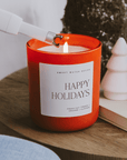 Lit Happy Holidays red jar candle styled beside white decorative Christmas trees