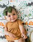 Baby in a mustard yellow outfit with a green headband sitting on a green blanket against a wall with a nature-themed wallpaper.
