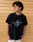 Man wearing the black Do You Believe in Jesus heavyweight tee beside a wooden wall.