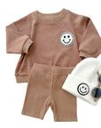 Children's brown outfit with smiley face design, white beanie, and sunglasses on a white background