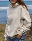Person wearing a white hoodie with text, standing by the ocean.