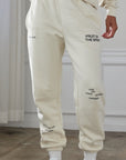 Person wearing a beige tracksuit with text on the pants, standing in a room with light-colored walls and wooden floor.