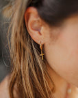 Angled close-up of a gold-plated cross hoop earring worn with hair tucked behind the ear
