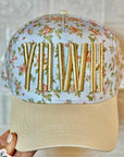 Floral-patterned cap with gold letters held by a hand, against a neutral background.