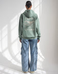 Back view of the moss green God Is Greater Hoodie showing embroidered lettering and a mountain-inspired design, styled with jeans.