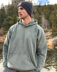 Man wearing the God Is Greater Moss Hoodie with a dark beanie outdoors.