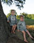 Two children seated beside a tree wearing Millstone Grey God Made Me hoodies and coordinating shorts