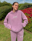 Woman wearing the Bordeaux God’s Promises Hoodie with a front kangaroo pocket outdoors beside flowering shrubs.
