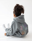 Seated child showing the floral back design on the God’s Still Working girls’ hoodie.