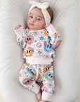 Baby wearing a colorful outfit with smiley faces and patterns, lying on a white cushion.