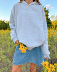 Jesus Is King Heather Grey Hoodie styled with a denim skirt in a field of yellow flowers