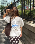 Woman wearing the Jesus Loves New York tee beside an iron fence in the city, showing the back design.