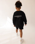 Child walking away in the Kingdom Club Jet Black crewneck sweatshirt with white Elevated Faith back lettering.