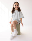 Child seated wearing the oversized Kingdom Club Open Air Blue Kids Tee with matching shorts