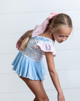 Young girl in a blue skirt and floral top standing on a wooden floor with a white wall background