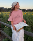 Model holding an open Bible while wearing the Ash Rose Love Your Neighbor tee with a white skirt