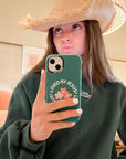 Woman wearing a straw cowboy hat and green sweatshirt holding the Loved by Jesus floral iPhone case for a mirror selfie