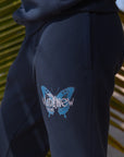 Black hoodie with a blue butterfly logo on the sleeve against a palm leaf background