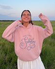 Model outdoors wearing the More Than Sunday Zephyr Pink Hoodie with blue script lettering