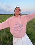 Model with arms outstretched wearing the More Than Sunday Zephyr Pink Hoodie outdoors
