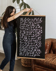Woman holding a black Psalm 23 poster with white script lettering and wooden hanging rails beside a brown leather chair.