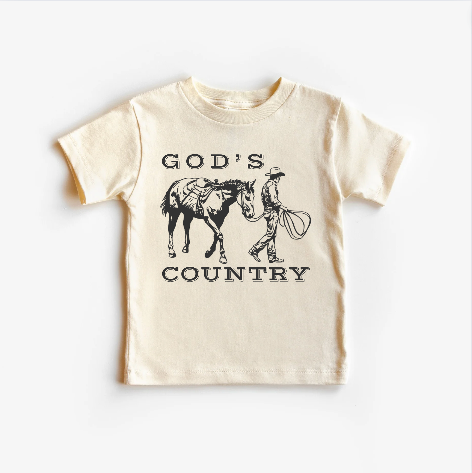 A beige neutral tee shirt top with 'GOD'S COUNTRY' printed in black text and a graphic of a horse and rider in black illustration on the front.