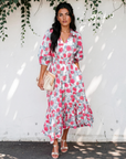 Model wearing a red and pink floral midi dress with puff sleeves and a tiered ruffle hem, styled with white heels and a beige handbag.