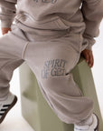 Child wearing a beige tracksuit with 'Spirit of G' text, sitting on a green stool.