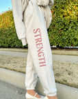 Person wearing light-colored sweatpants with 'STRENGTH' printed on them, standing outdoors.