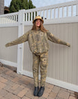Woman wearing The Sacrifice Forest Camo sweatpants with a coordinating hoodie beside a light-colored fence.