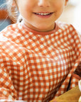 Young girl wearing a red and white checkered shirt, sitting at a table with a blurred background.